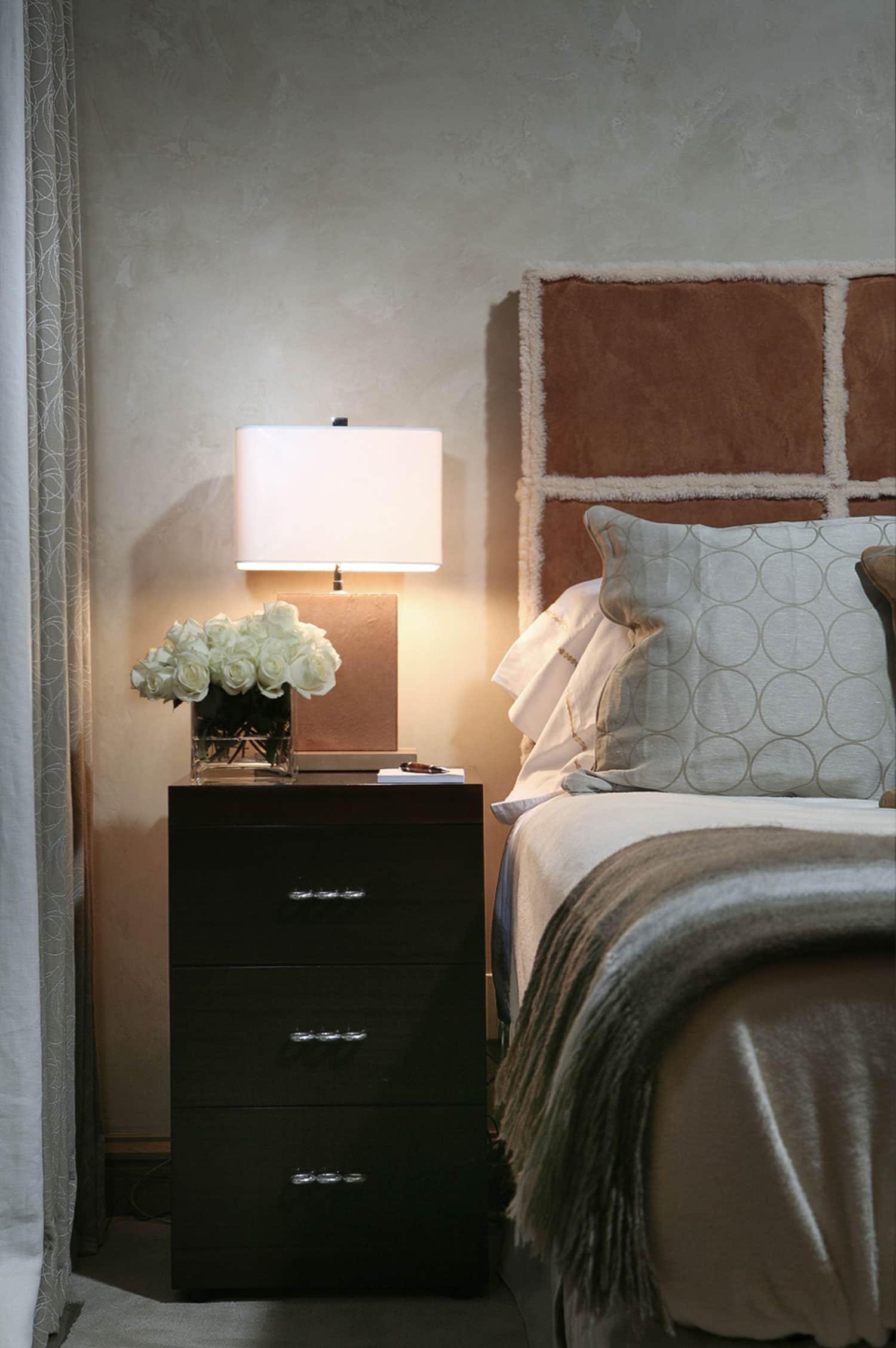 In this Aspen residence designed by Carol Egan, this image is a detail of the Master bedroom showing a custom grided shearling headboard with Dunbar wooden three drawer bedside table and a rectangular ceramic camel colored glazed table lamp.