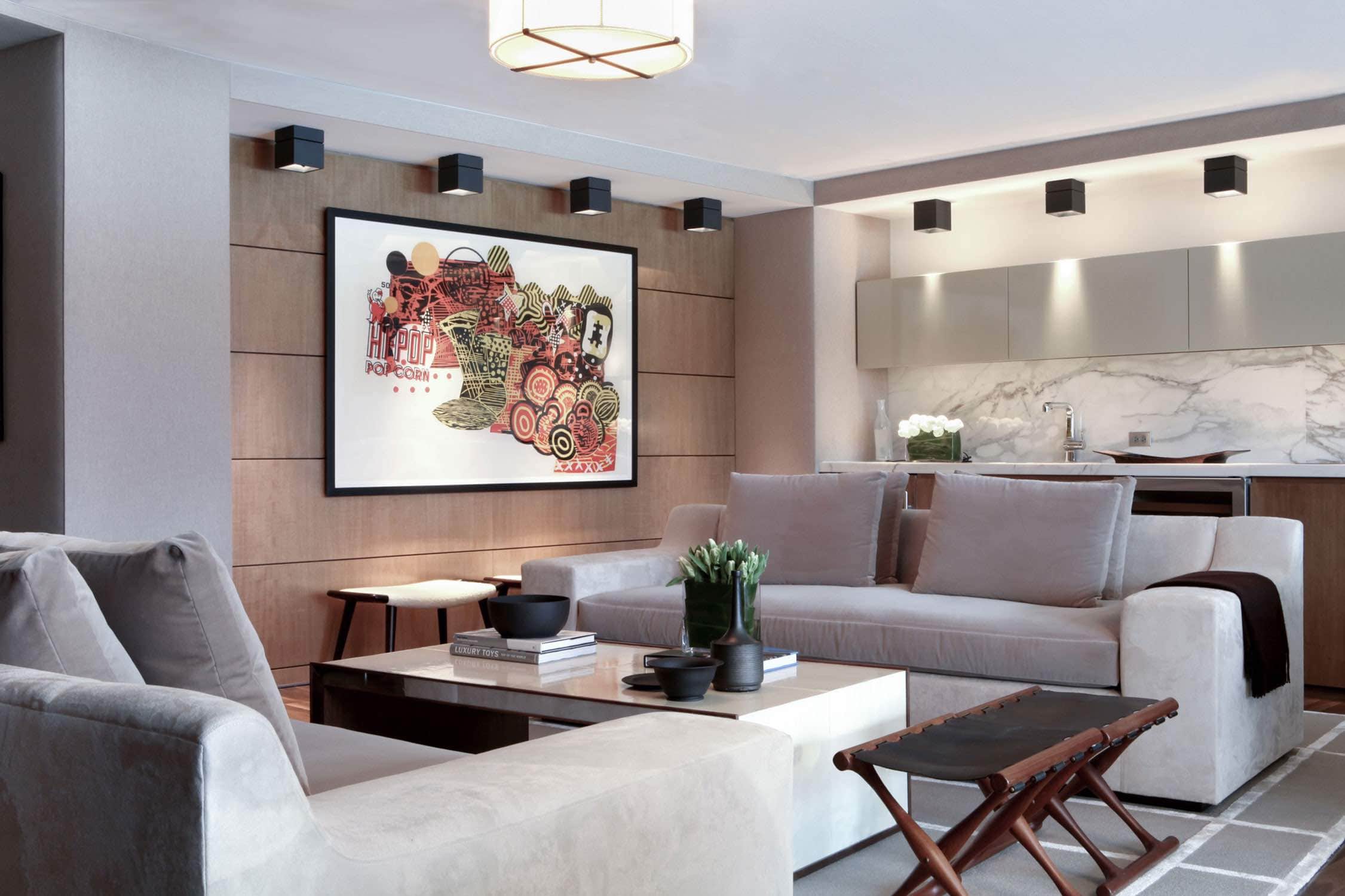 This image shows the owners viewing suite at the Citifield ballpark.  Upholstered and oak paneled walls border a built in Kitchen and Bar area.  Hand woven area rug with two suede JMF style sofas surround a “Samuel" coffee table by Roman Thomas. There is a parchment lampshade ceiling fixture by Roman Thomas and two Scandinavian folding leather and walnut benches in the foreground.