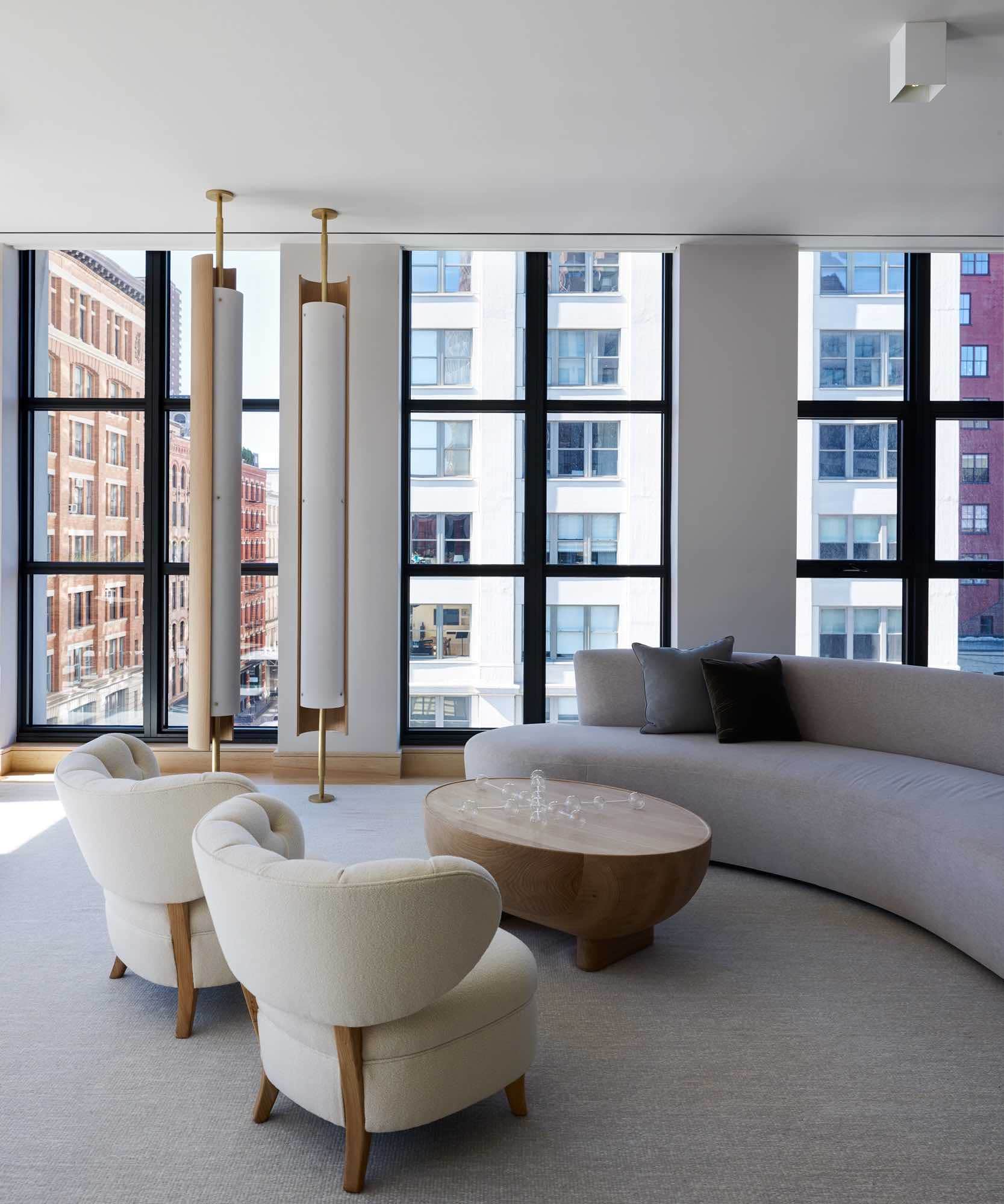 This image shows the living room area in the open plan loft layout in the Tribeca apartment designed by Carol Egan Interiors.  A serpentine shaped custom sofa design by Studio Carol Egan sits opposite a pair of Vintage lounge chairs by Otto Schulz with a sculptural Oak coffee table.  There are two Oak and Brass Pillar lights floor to ceiling designed by Studio Carol Egan and a Hand-woven wool area rug in heathered grey tones while the upholstery of the sofa and chairs and neutral boucles and textural neutral mohairs.