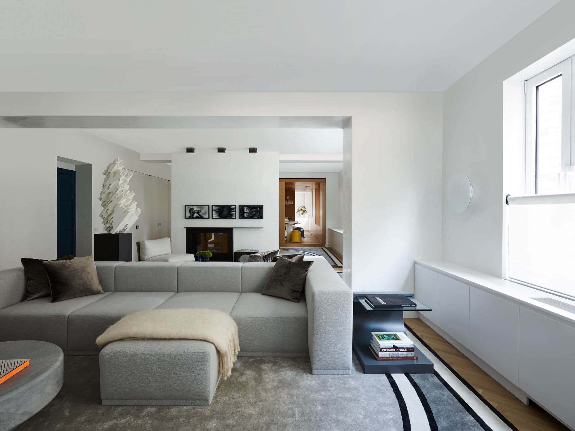 In this living room designed by Carol Egan, the two-sided sofa is a modular custom designed sectional sofa accessible from the north and south sides in the living room.  One side orientates towards the fireplace, the other side orientates to the television viewing area.  The sofa is anchored by the graphic Roman carpet design by Eric Schmitt Studio.  There is a custom designed circular coffee table in Bleu de Savoy stone along with a sofa cantilever side table by Karl Springer.