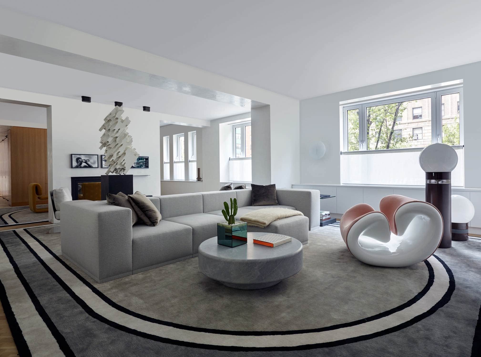 Designed by Carol Egan, this image shows a wide-angle view into the living room of a Park Avenue Apartment.  In the foreground, the sculptural and playful Esox chair by jean Pierre Laporte is upholstered in a powder Pink Mohair with White fiberglass frame.   This composition is completed with Orocolo floor lamps by Gae Aulenti sitting on the floor next to the chair.  The graphic black and white and grey Roman Area rug by Eric Schmitt creates a racetrack feeling to the open plan loft layout.  A double sided custom modular sofa by Studio Carol Egan is upholstered in a grey boucle fabric called Brunswick Feather by Holland & Sherry.