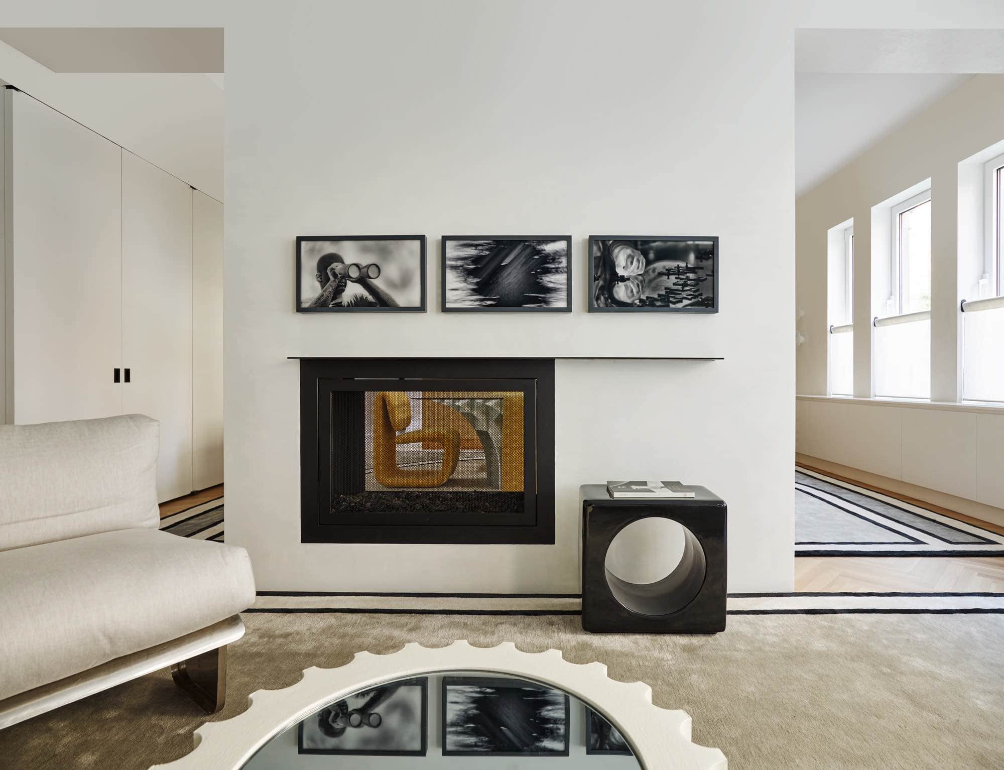 This photograph shows a detail of a 1970s Chauffeuse stainless steel chair upholstered in a pale grey linen fabric and positioned by the fireside in the living room of this Park Avenue Apartment designed by Carol Egan.  In the foreground is a detail of the mirror reflection in the Guffram Attica TL coffee table by Studio 65.  Above the fireplace there are three framed Gelatin print photographs by Richard Mosse from the "incoming" Series.  A ceramic Ringo side table by India Mahdavi is placed by the fireside.