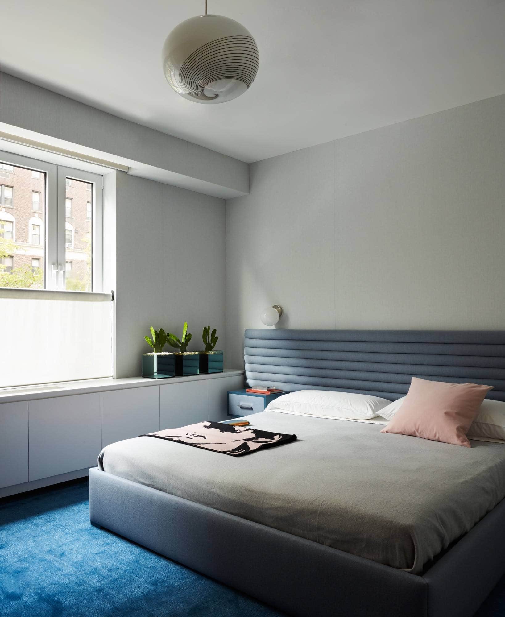 Shades of blue define this Park Avenue bedroom designed by Carol Egan.  Seen in this image are pale blue upholstered walls with a rolled contrast blue Flannel headboard that is wall to wall in the room.  A navy blue and Prussian Blue Nepalese silk carpet is fitted wall to wall in the master suite and dressing room area.  On the ceiling is a 1970s Murano glass globe pendant by Gino Vistosi. Tip of the Tongue wall lights by Michael Anastassiades flank the bedside tables which are integrated in the upholstered headboard.  Dennis Hopper portrait by Andy Warhol printed on Wool Saddle Blanket accessorizing the bed.