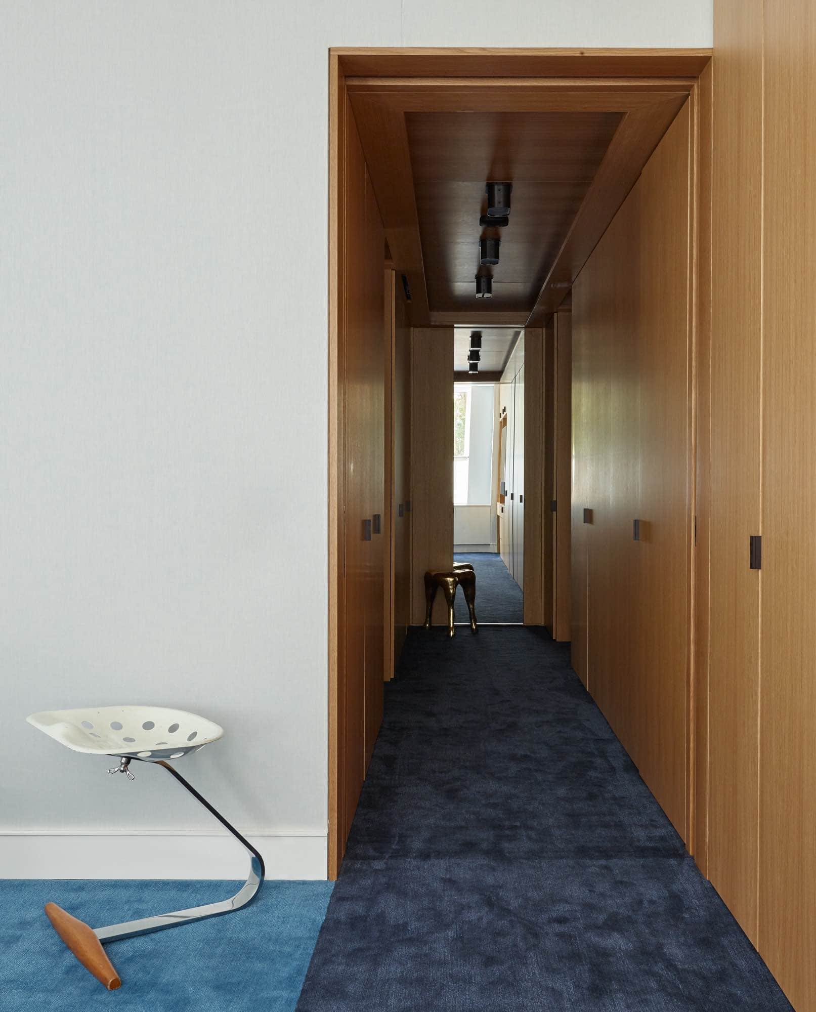 "Mezzadro" stool by Achille & Pier Giacomo Castiglioni is shown in this photograph next to a pale blue linen upholstered bedroom wall with door frames and dressing room millwork in Oak finish in this Master bedroom and Dressing Room Suite designed by Carol Egan.  Custom twill carpet by Carini Lang in navy and Prussian Blue contrasting colors.