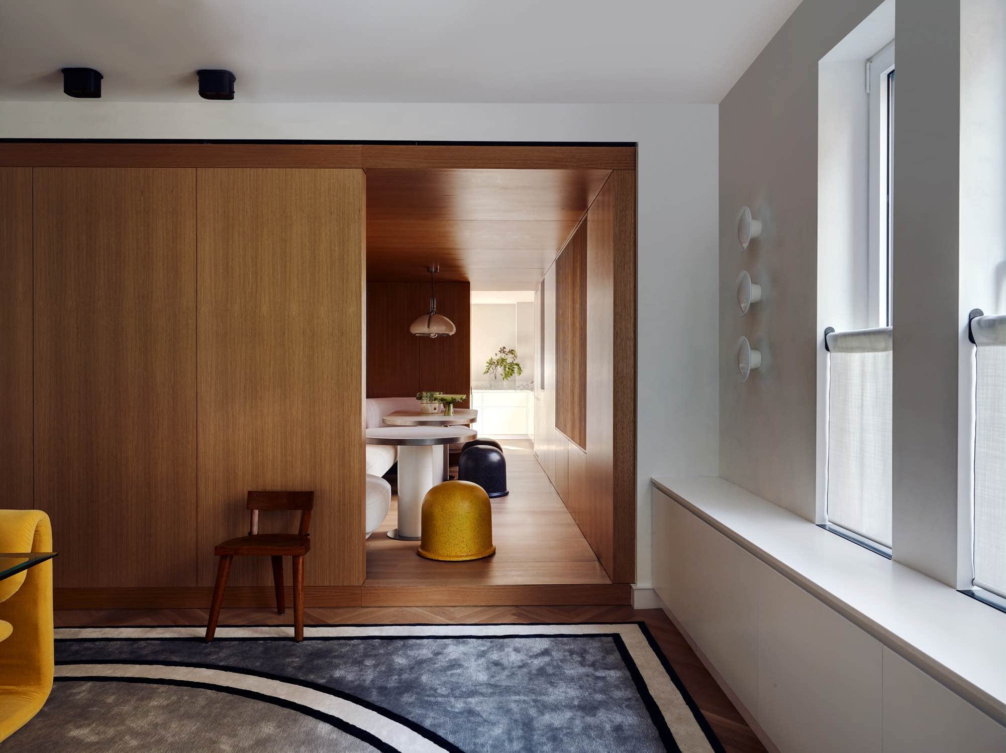 Designed by Carol Egan, this image is a view through the dining room into the oak paneled breakfast room.  The Roman carpet design by Eric Schmitt Studio can been seen in the dining room in black border outline with grey tones creating the racetrack design in the rug.  Puffo stool by Gruppo Sturm can be seen flanking the dining table in the breakfast room.  A Quadrifoglio pendant light by Gae Aulenti hangs above the dining table.  Three AJ Eklipta by Arne Jacobsen wall lights are stacked on the dining room wall.
