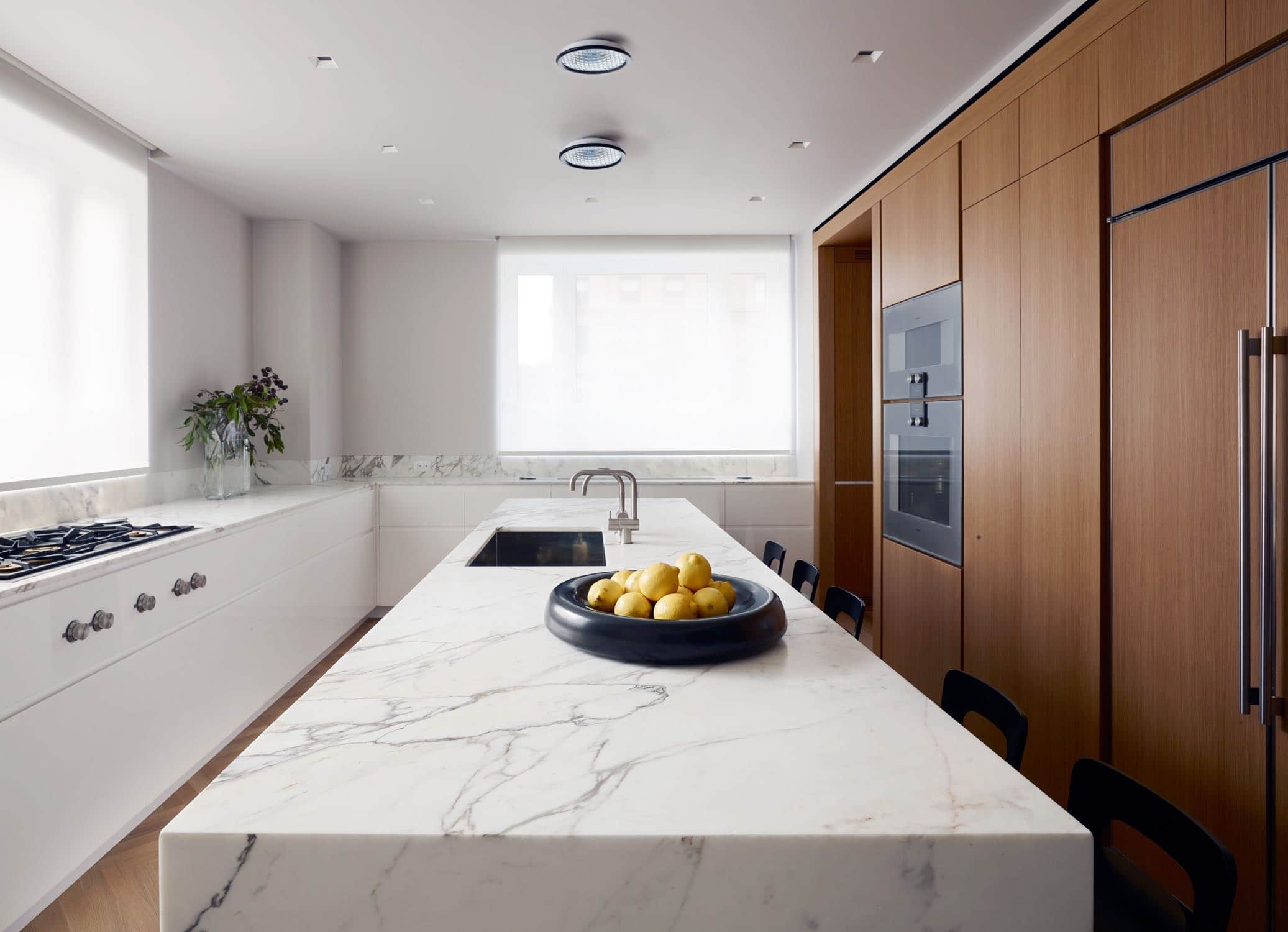 1960s flush mount lights by Stilnovo are seen in this image on the kitchen ceiling over the marble island.  The kitchen is comprised of a wall of full height oak cabinetry, a Statuary honed marble island and the perimeter of the kitchen has lower cabinets in white painted finish with windows above giving double exposure light to the room.  K65 highchairs in ebony finish by Alvar Aalto are seen tucked under the statuary marble kitchen island.  One handle mixer faucet by Vola.