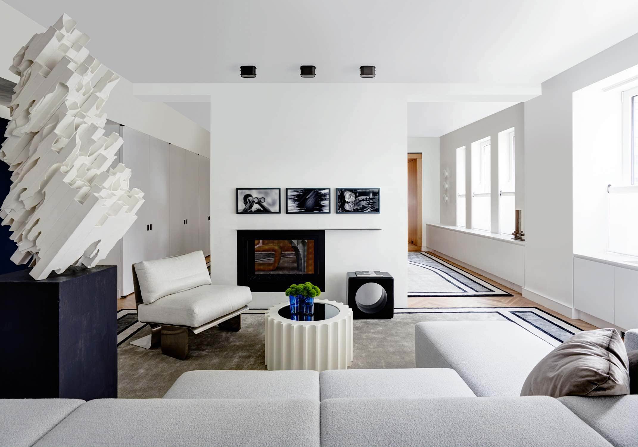 A 1970s Chauffeuse stainless steel chair flanks the fireplace in this image of the living room of a park avenue apartment designed by Carol Egan.  A Custom modular sofa upholstered in Brunswick Feather fabric by Holland & Sherry is shown next to the Guffram Attica TL coffee table by Studio 65.  The ceramic Ringo side table by India Mahdavi can be seen next to the fireplace. Lightning Sculpture by Kouzo Takeuchi is seen in the foreground on a wooden pedestal. Artwork from the Incoming Series h Photography by Richard Mosse hangs above the fireplace. Roman carpet design by Eric Schmitt Studio.