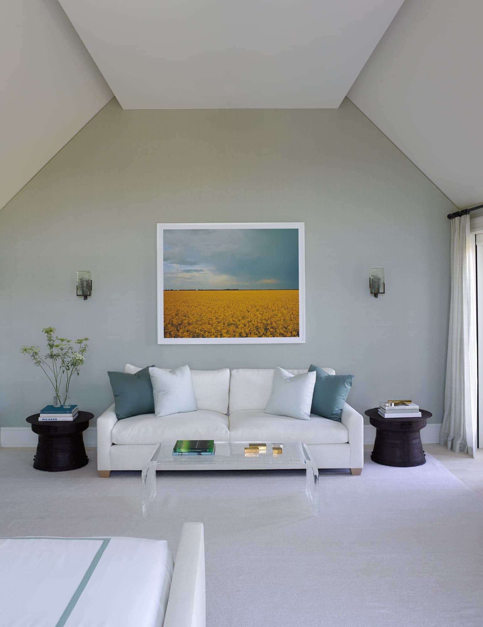 Designed by Carol Egan, this image shows a master bedroom with upholstered walls in custom "Jed’s Pale Blue Silk" fabric by Sam Kasten.  There is a JMF style sofa upholstered in white linen and flanked by African drum tables in blackened metal.  The wall lights are glass by Lianne Gold and they hang alongside a photograph by Larysa Sendich.