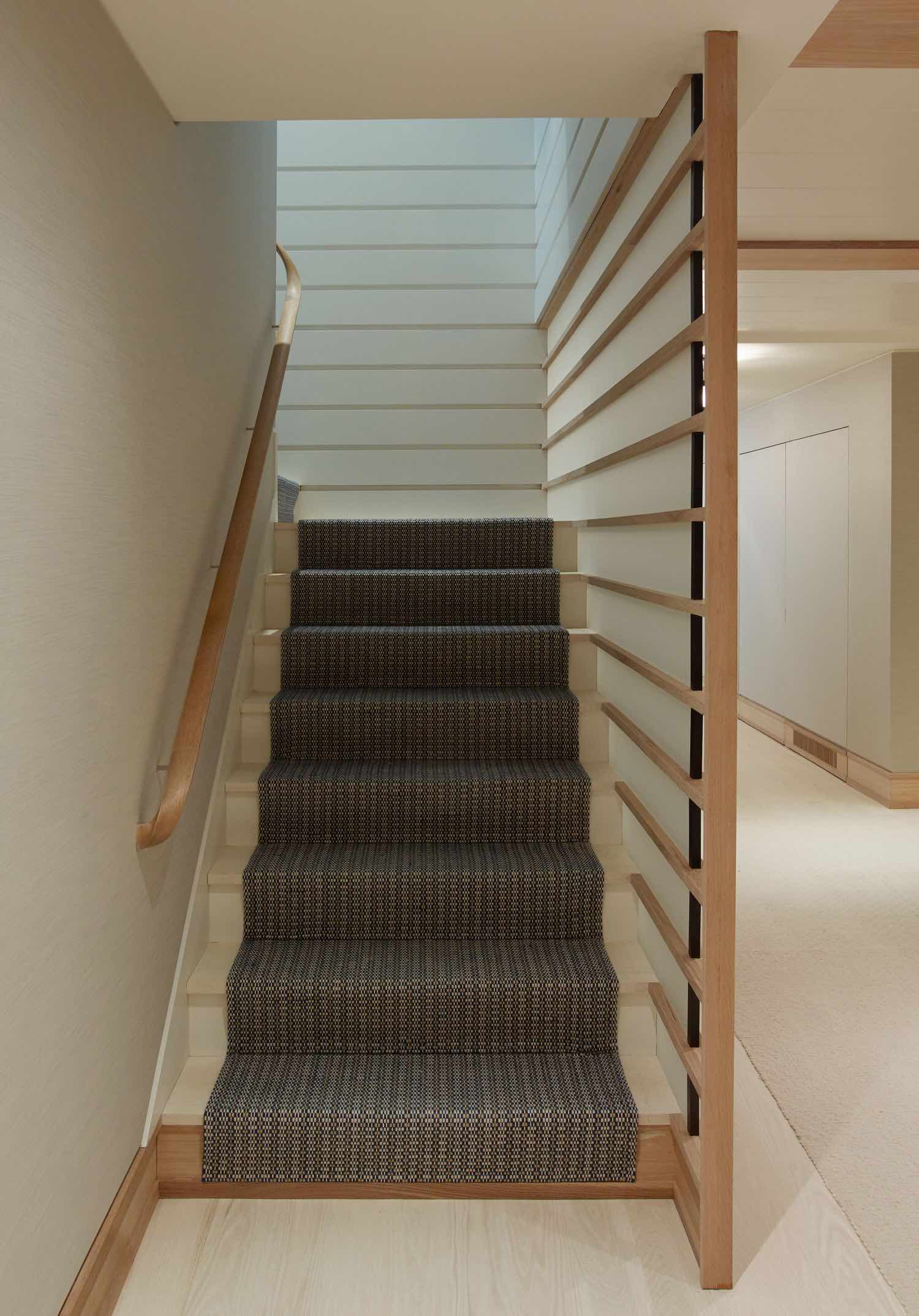 Designed by Carol Egan, this image shows a staircase that is flanked by a custom hand-carved white oak handrail and oak and etched glass wall.  The oak staircase is fitted with a custom woven Elizabeth Eakins woven wool runner in navy blue color.
