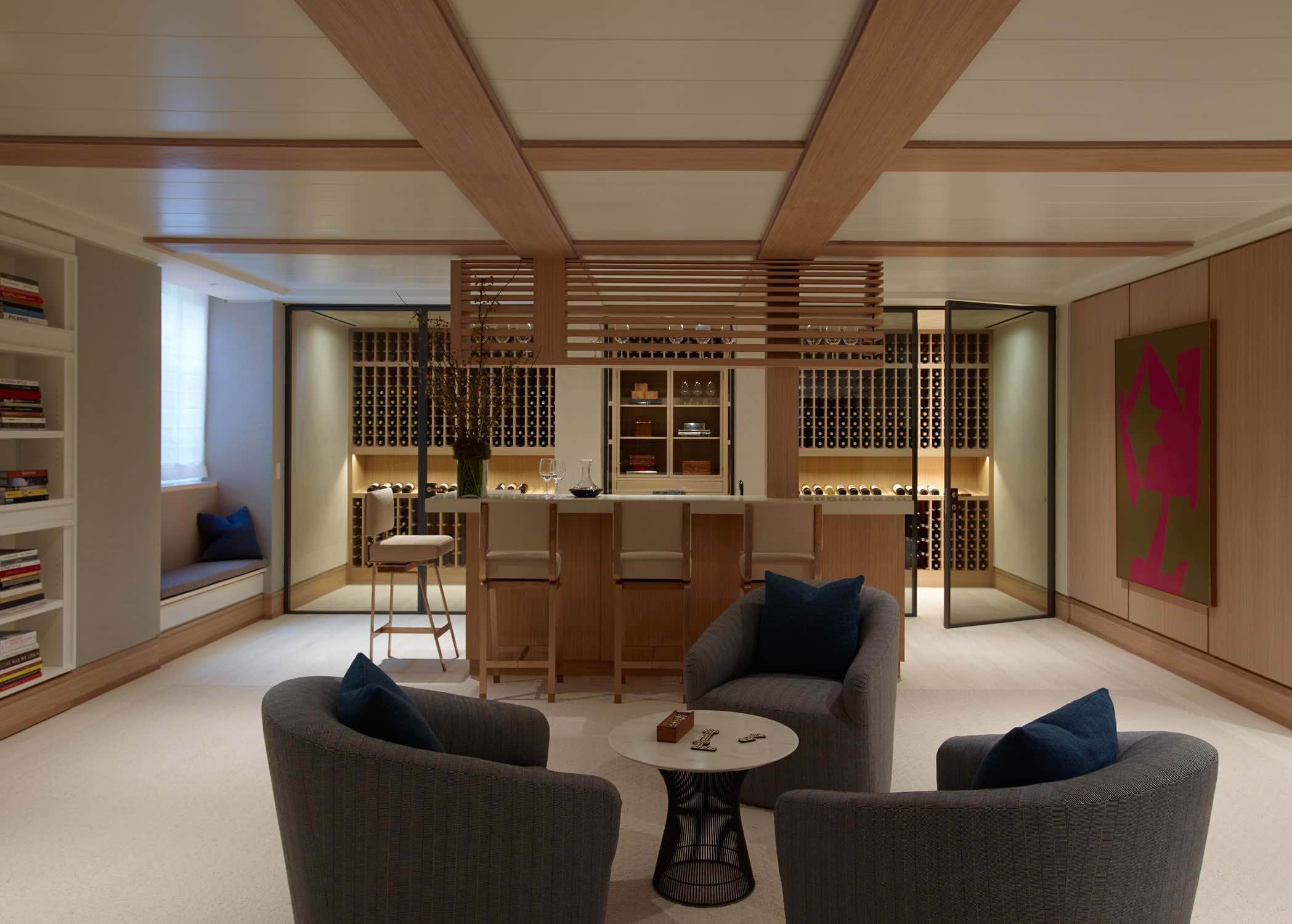 This image shows a Custom Wine Cellar by Studio Carol Egan in oak with bronze framed glass doors and oak paneled walls.  A large painting by Jorge Fick is hung outside the cellar and there is a bar area with glass storage hanging from the paneled ceiling with custom metal and upholstered bar stools from Khouri Guzman Bunce.