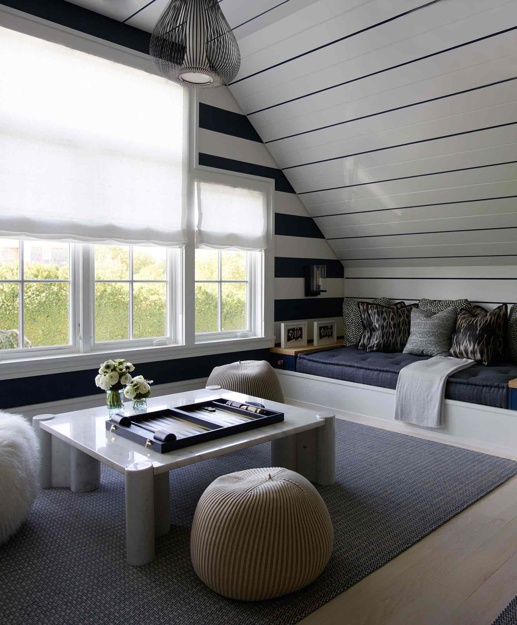 This image shows a detail of the nook at the top of the stairs designed by Carol Egan.  The ceiling is clad in painted V grove board with an indigo blue painted stripe.  A wall light cylinder sconce by Comerford Collection lights the nook.  There is an area rug in deep blue by Elizabeth Eakins.  Build in banquettes share a large stone coffee table with Bonnet poufs by Casalis around the Jumbo coffee table by Gae Aulenti.