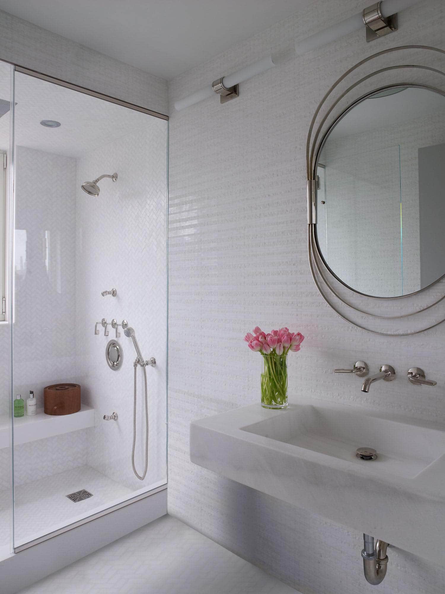 This image shows a bathroom designed by Carol Egan with walls and floors made of chisel cut Thassos marble.  The vanity sink is fabricated from slab Thasos marble and hand carved.  Plumbing fixtures and fittings are the .25 range in polished nickel finish from Waterworks.  A Vintage Nickle framed mid-century mirror hangs above the sink.   The room is lit with a 1950s antique glass ceiling fixture from Valerie Goodman Galerie and Opus wall lights from Waterworks.